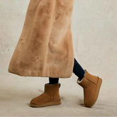 Person wearing brown fur-lined suede boots and a long brown coat on a neutral background